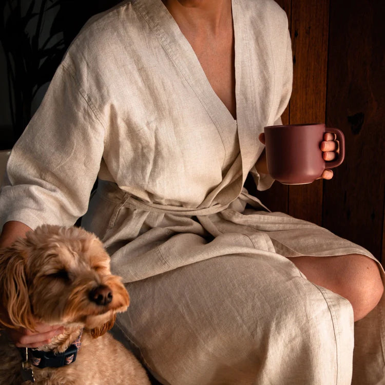 Woman wearing natural european flax linen robe holding a coffee cup, patting a dog, watching the sunrise